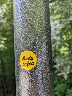 A yellow circular sticker with the text "Rudy cafea" in a stylized script font. It's affixed to a textured gray metal pole outdoors, with a natural green background visible.