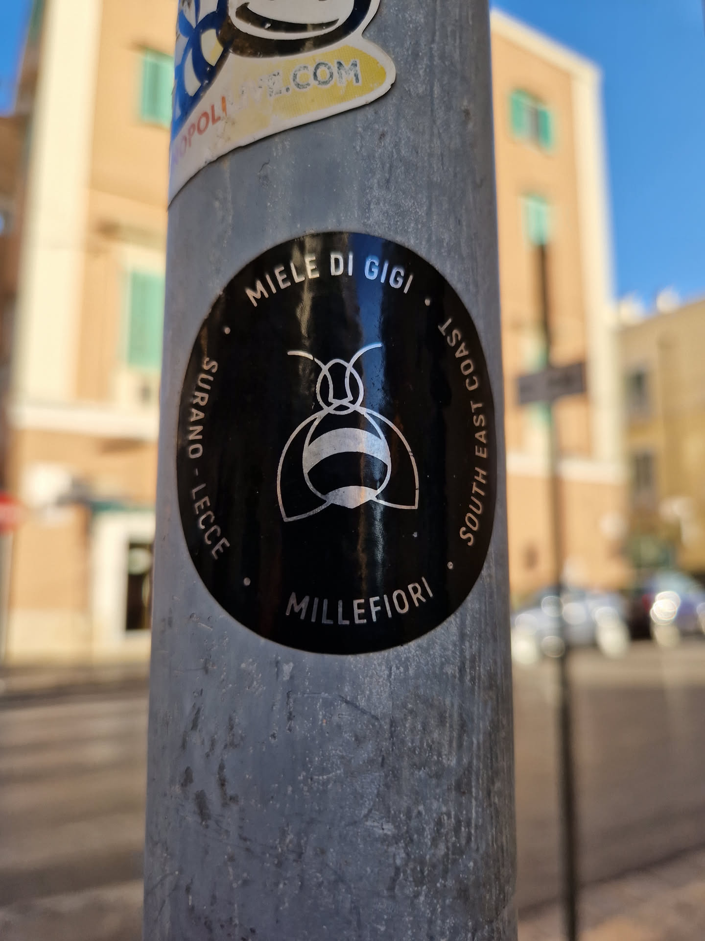A black and white round sticker with a bee illustration in the center. The text around the bee says "Miele di Gigi", "Surano - Lecce", and "South East Coast". At the bottom it says "Millefiori".