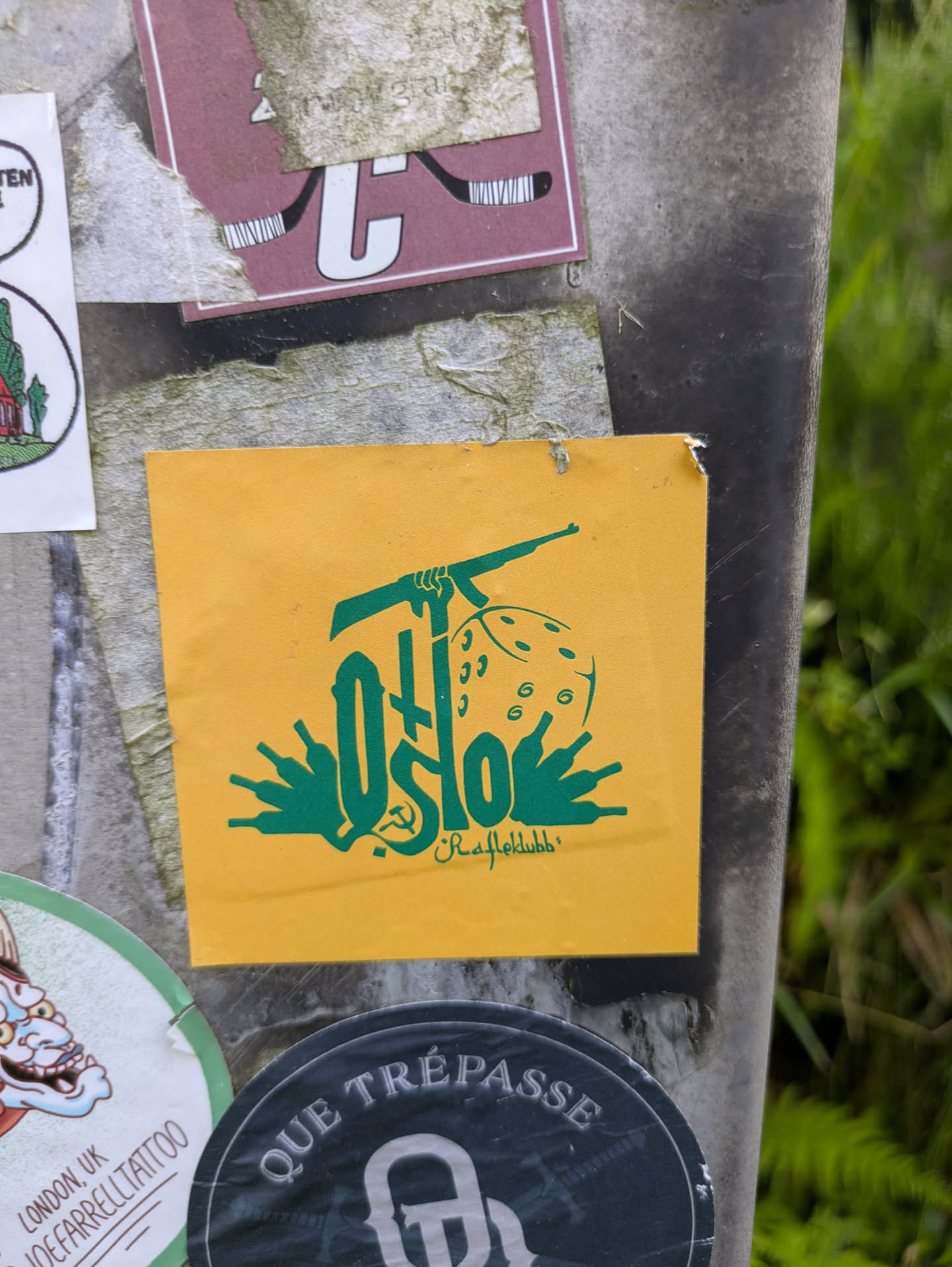 The central sticker is a square, vibrant yellow design with all elements rendered in green. The dominant text is 'Oslo', with the 'O' creatively forming a hand gripping what appears to be a rifle. To the right, a six-sided die is visible, and below the word 'Oslo', several stylized bottle shapes are depicted. A small hammer and sickle emblem is subtly integrated near the base of the 'o'. Beneath the main graphic, in a smaller, cursive font, the word 'Ræfleklubb' is written. The combination of these symbols suggests a commentary that could be political, social, or related to a specific subculture in Oslo, Norway.