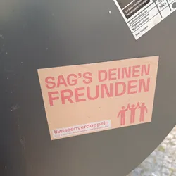 A beige sticker with the text "SAG'S DEINEN FREUNDEN" (Tell your friends) in pink letters.  Below the text are three figures giving each other a high-five.  A small hashtag and website are visible at the bottom. 