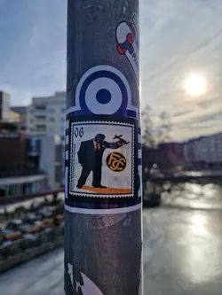 This sticker has a retro design, appearing as a postage stamp.  The central image depicts a man in a suit holding a scale with a circular emblem that has the letters FC. The colors are mainly black, orange, and white. The sticker is applied to a metal pole. No QR code or social media links are visible.