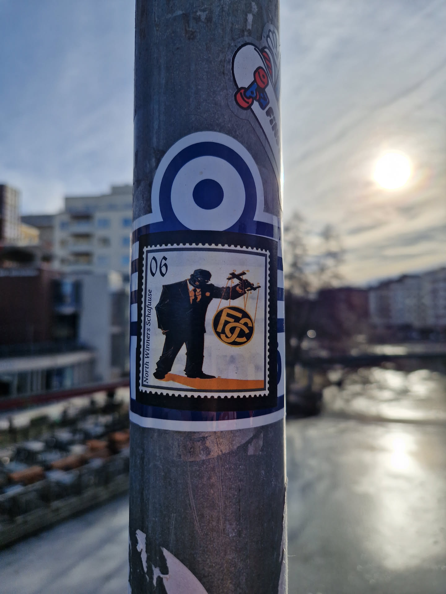 This sticker has a retro design, appearing as a postage stamp.  The central image depicts a man in a suit holding a scale with a circular emblem that has the letters FC. The colors are mainly black, orange, and white. The sticker is applied to a metal pole. No QR code or social media links are visible.