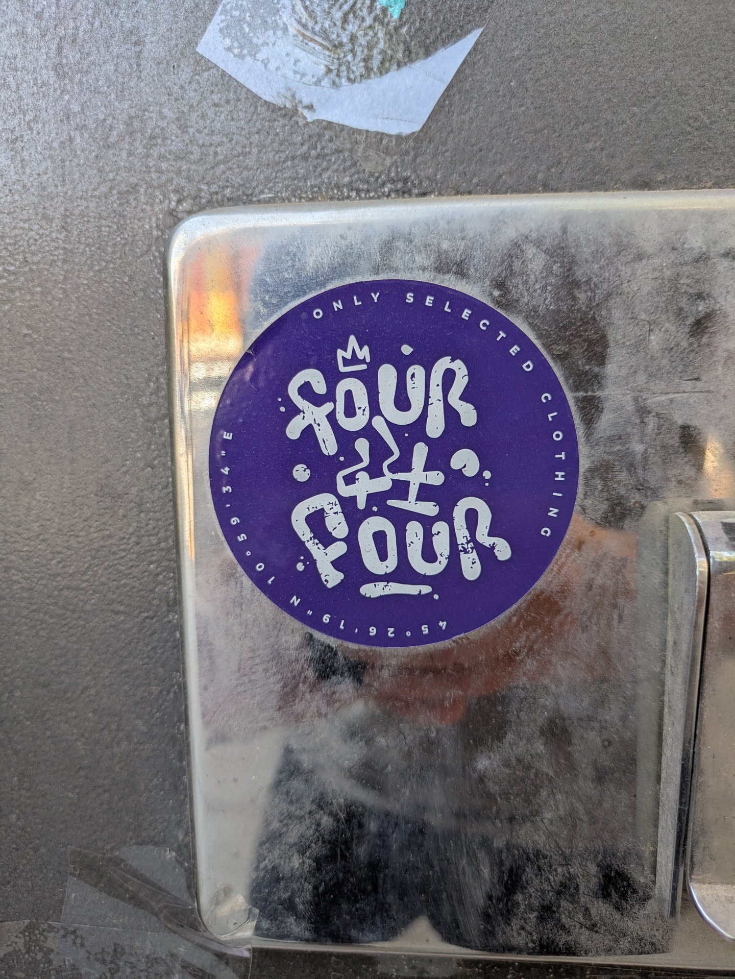 This is a round purple sticker with a white stylized design. The text "FOUR FOUR" is stacked in the center, with a small crown above the first "FOUR". The text "ONLY SELECTED CLOTHING" curves around the top edge of the sticker, and coordinates "45°26'19" N 10°59'34" E" are at the bottom.