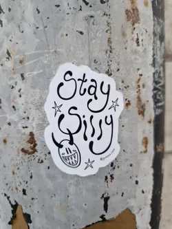 Street sticker A white sticker with black ink. It says Stay Sissy in a stylized font. There's a small devil-like face with horns and a toothy grin under the text, along with small stars on either side. The sticker is affixed to a weathered gray metal surface.
