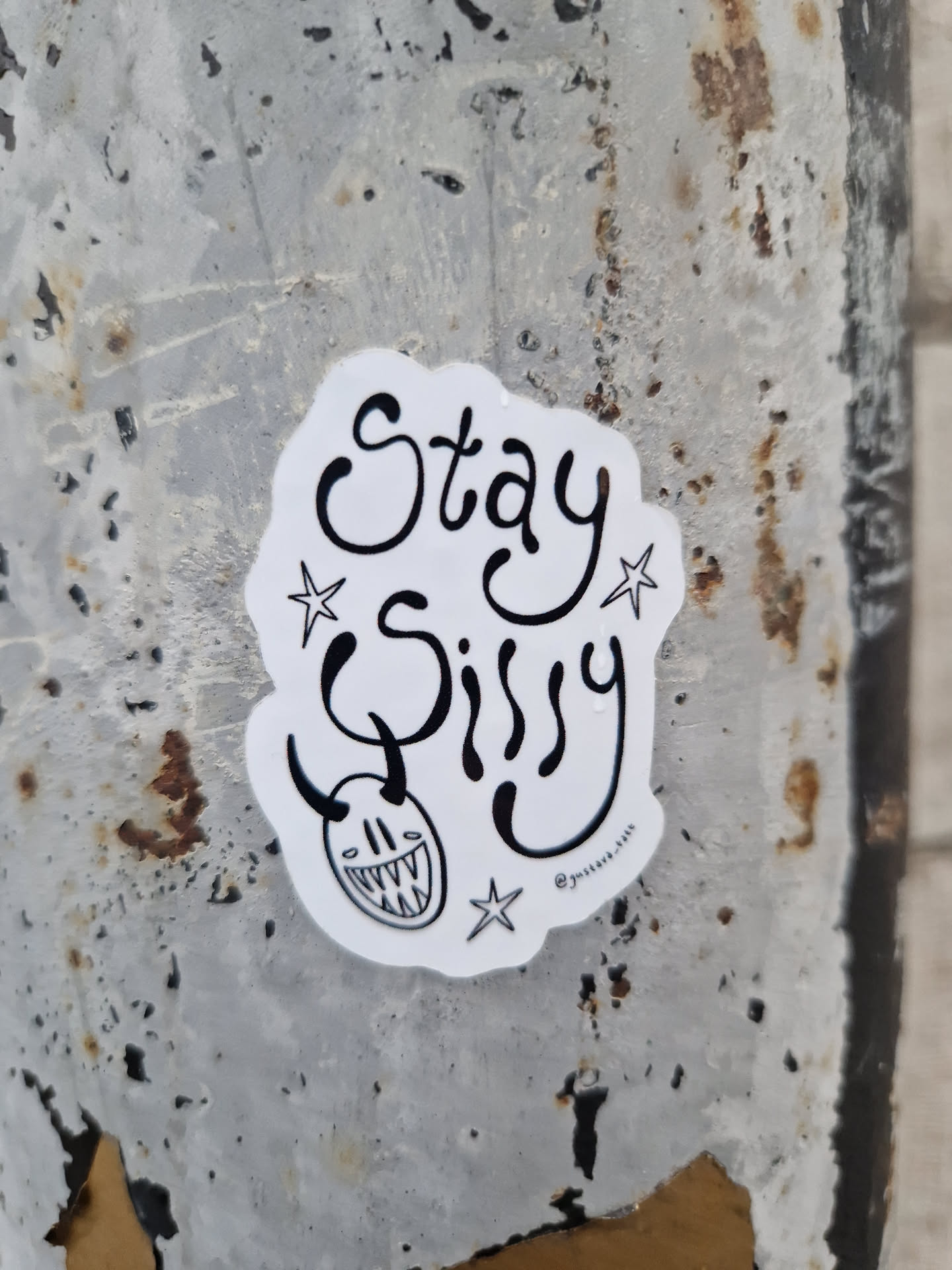 A white sticker with black ink. It says Stay Sissy in a stylized font. There's a small devil-like face with horns and a toothy grin under the text, along with small stars on either side. The sticker is affixed to a weathered gray metal surface.