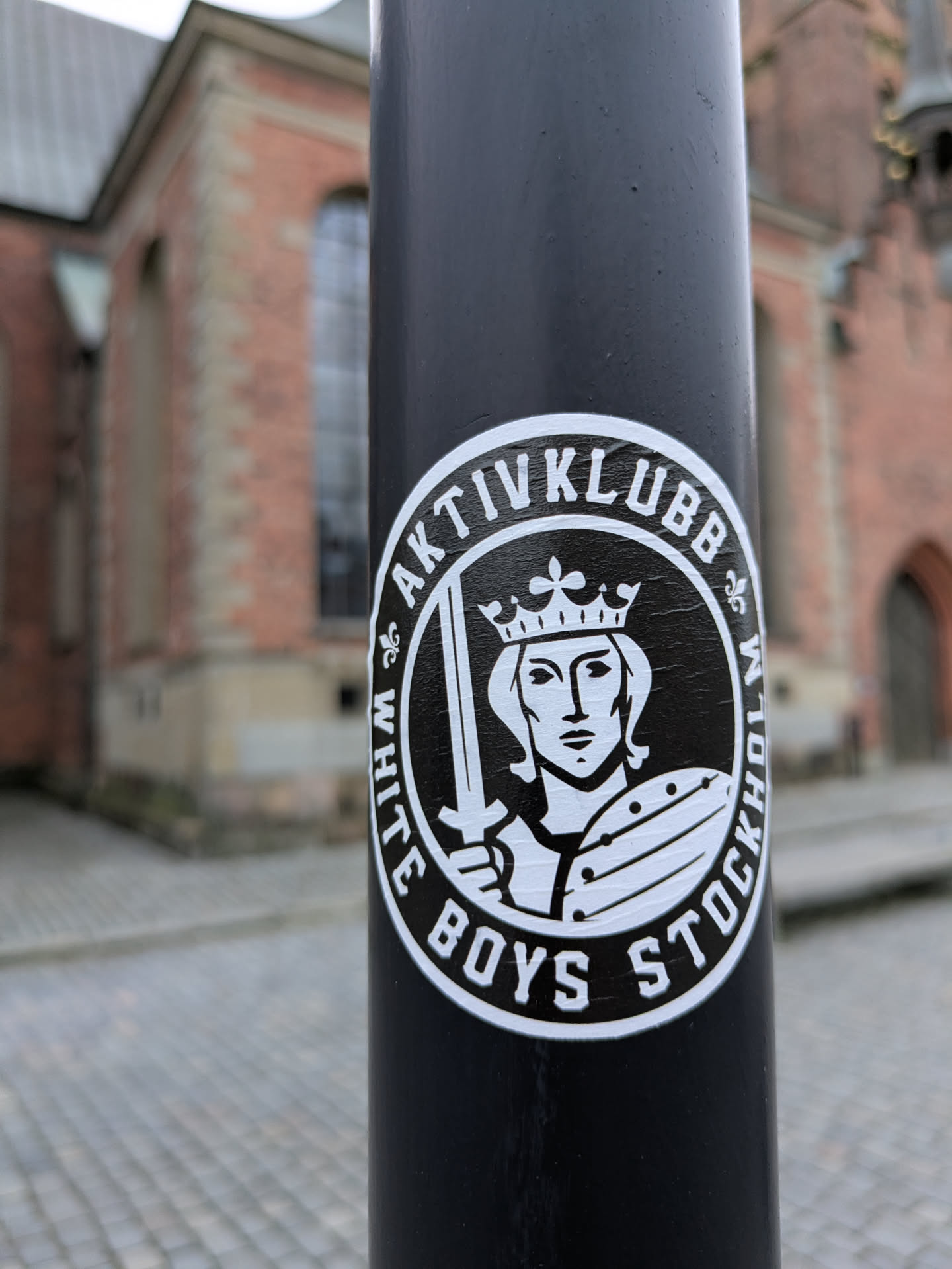 Black and white sticker with the text "White Boys Stockholm Aktiv Klubb" in a circular design. It features an illustration of a crowned figure holding a sword.