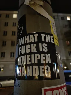 A black and white sticker on a pole with the text "What the f*ck is Kneipenbande?" in bold white letters on a black background. The sticker has a slightly worn look, and there are other stickers on the pole.