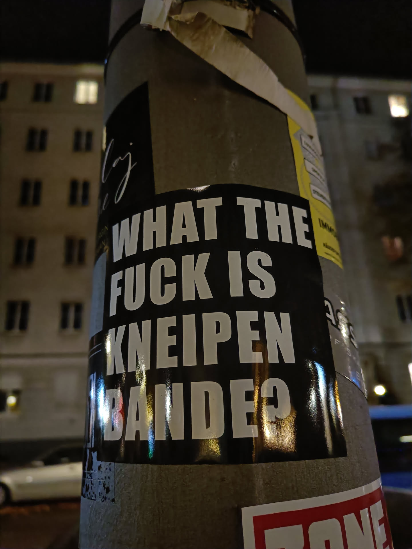 A black and white sticker on a pole with the text "What the f*ck is Kneipenbande?" in bold white letters on a black background. The sticker has a slightly worn look, and there are other stickers on the pole.
