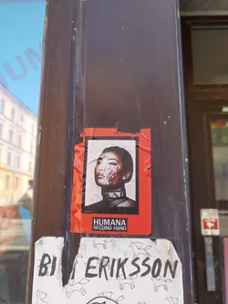 A vibrant red sticker with a close-up image of a person's face with painted details.  The text 'HUMANA SECOND HAND' is visible at the bottom. The sticker appears to be part of a campaign for the secondhand clothing brand, Humana.