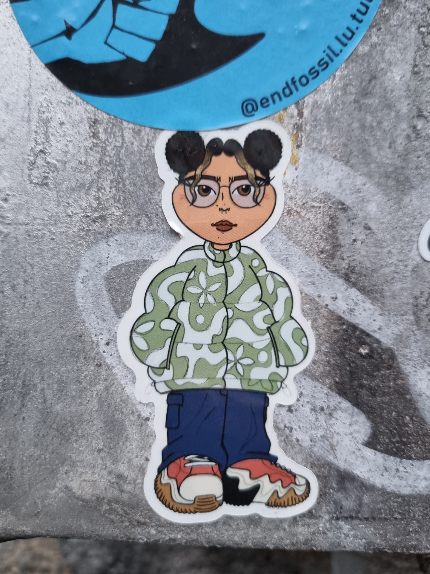 A sticker featuring a cartoon drawing of a girl with brown skin, glasses, and her hair in two buns. She's wearing a light green jacket with a pattern, dark blue pants, and red and white sneakers. The art style is reminiscent of a chibi or similar cartoonish character.