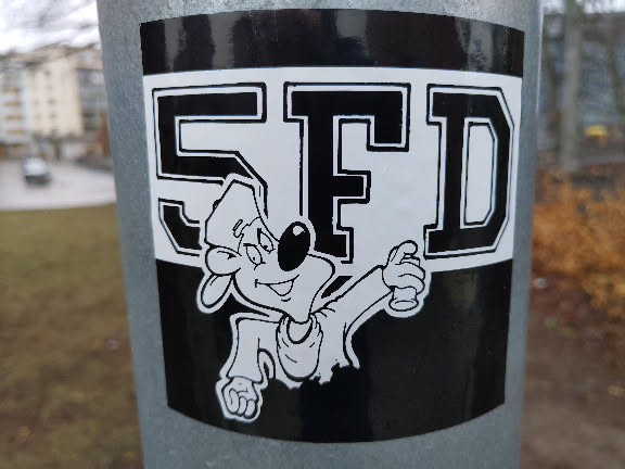 Black and white sticker featuring a cartoon character holding a spray can, with the letters SFD prominently displayed.  The style is reminiscent of 90s hip-hop and graffiti culture. The sticker is affixed to a metal pole outdoors.