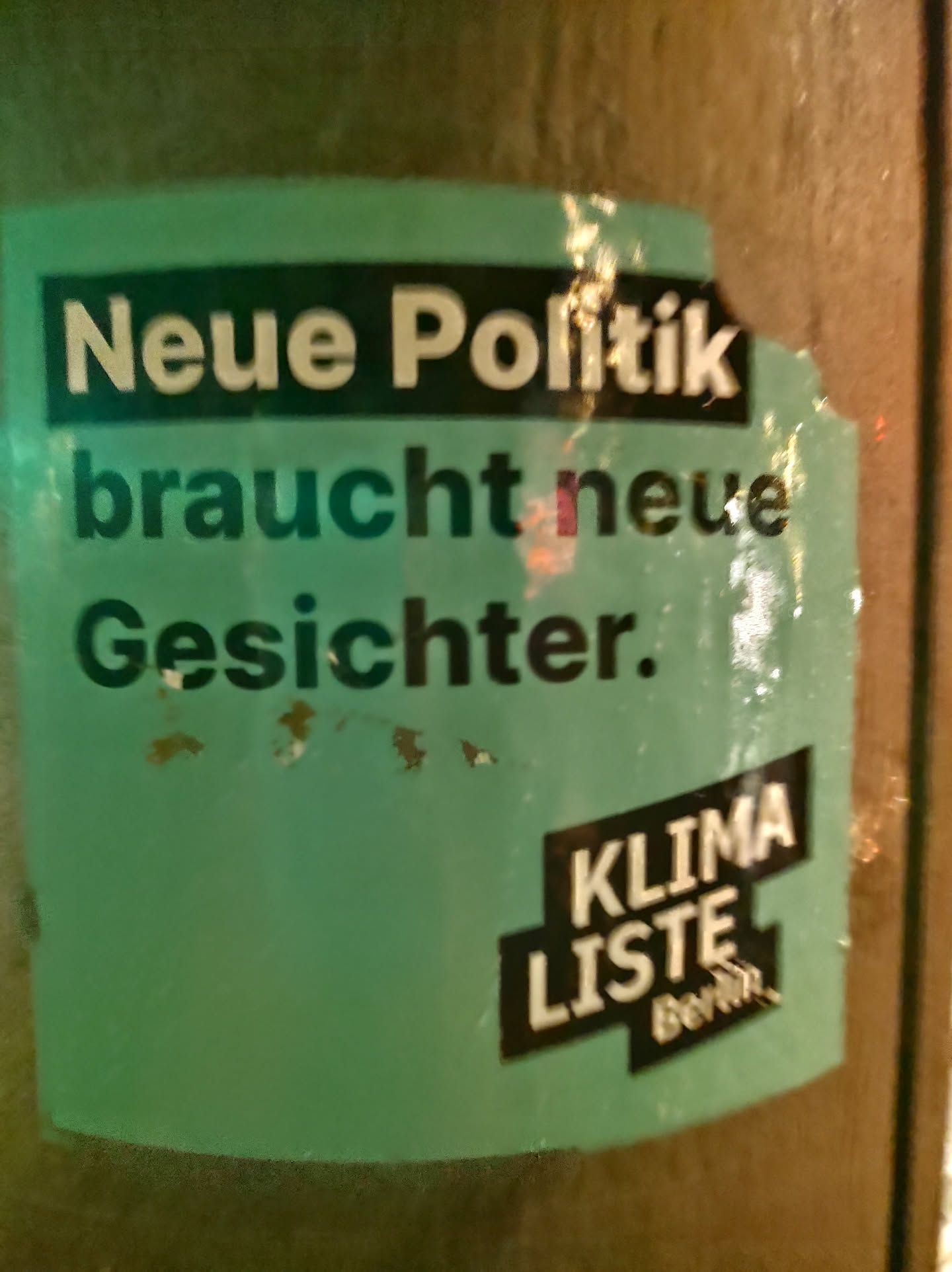 A teal and black sticker with the text "Neue Politik braucht neue Gesichter."  in white. At the bottom, it shows "KLIMA LISTE Berlin".