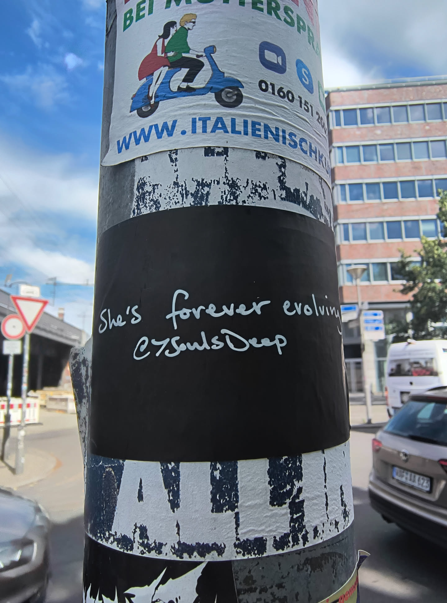 The sticker is black with white handwritten text. The text appears to be a quote followed by a social media handle. The edges of the sticker are visible, and it is applied to a pole that has other posters and peeling paper adhered to it. The writing is in a flowing, cursive font.