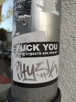 A black and white sticker with a provocative message. The sticker is attached to a metal pole and shows some wear and tear. It has graffiti-style text at the bottom.