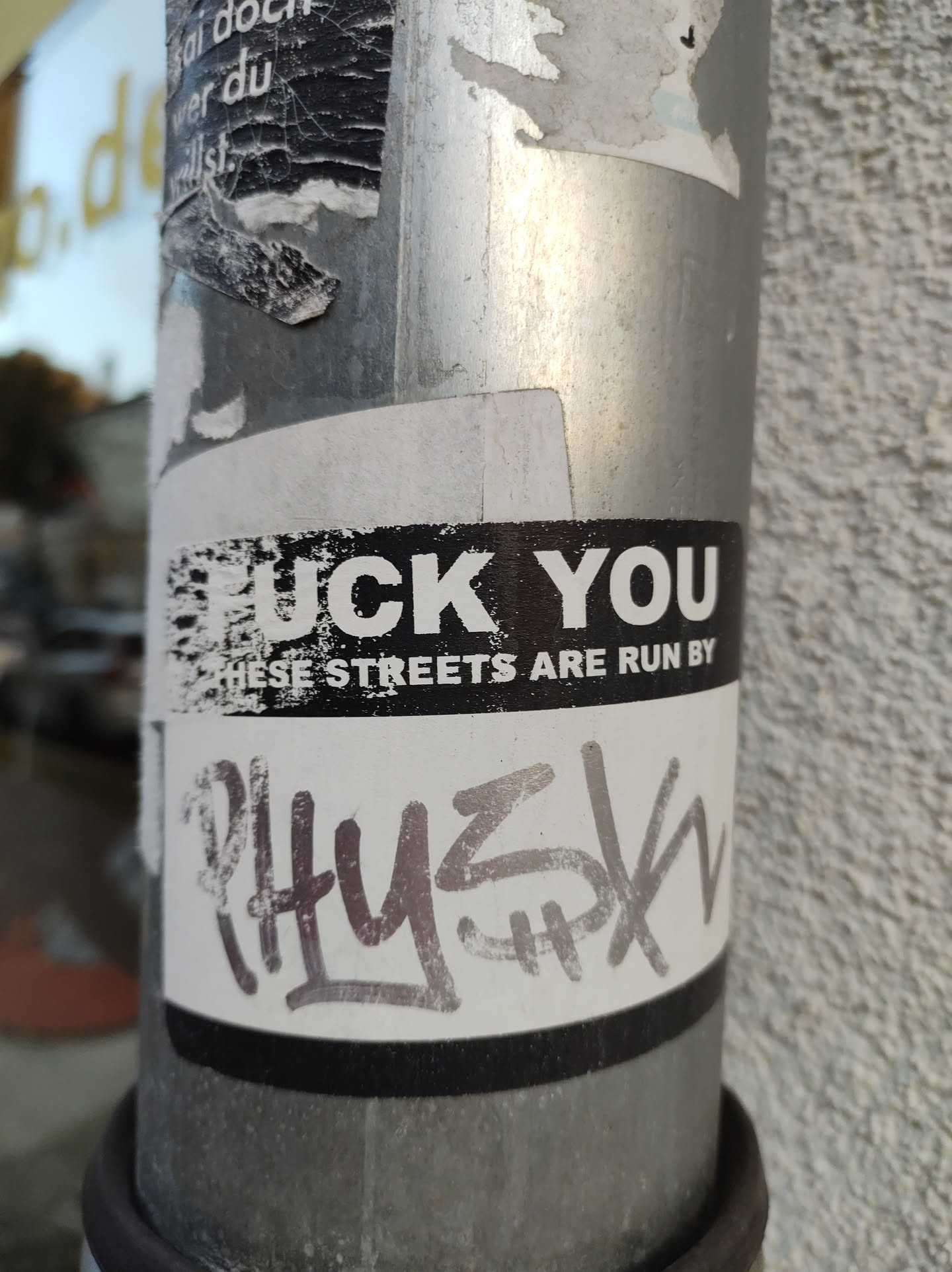 A black and white sticker with a provocative message. The sticker is attached to a metal pole and shows some wear and tear. It has graffiti-style text at the bottom.