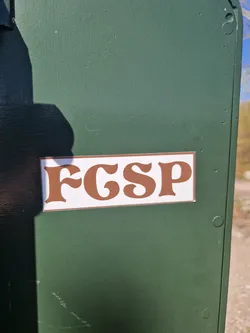 A brown and white sticker with the letters FGSP in a retro font, affixed to a dark green surface. 