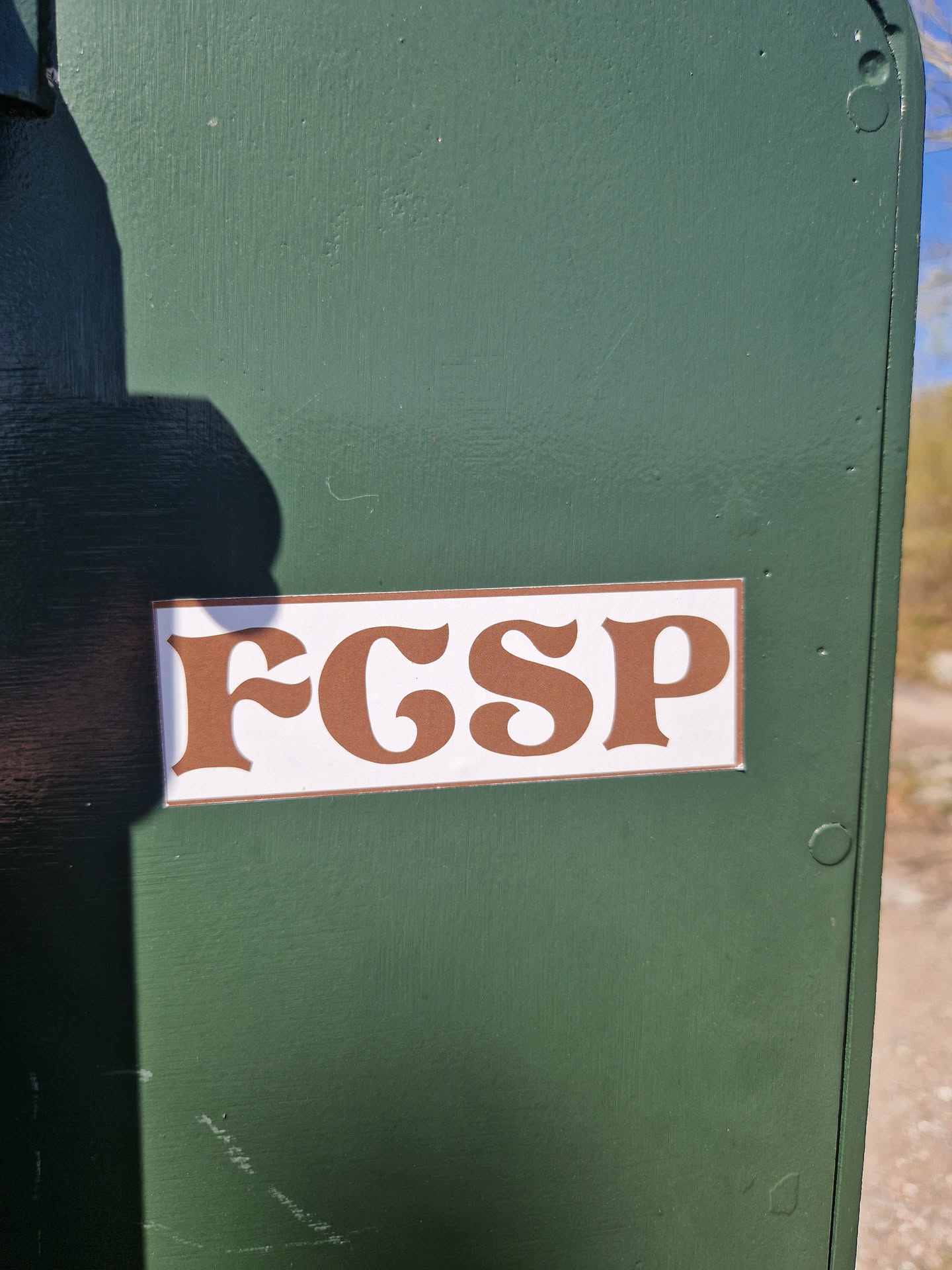A brown and white sticker with the letters FGSP in a retro font, affixed to a dark green surface. 