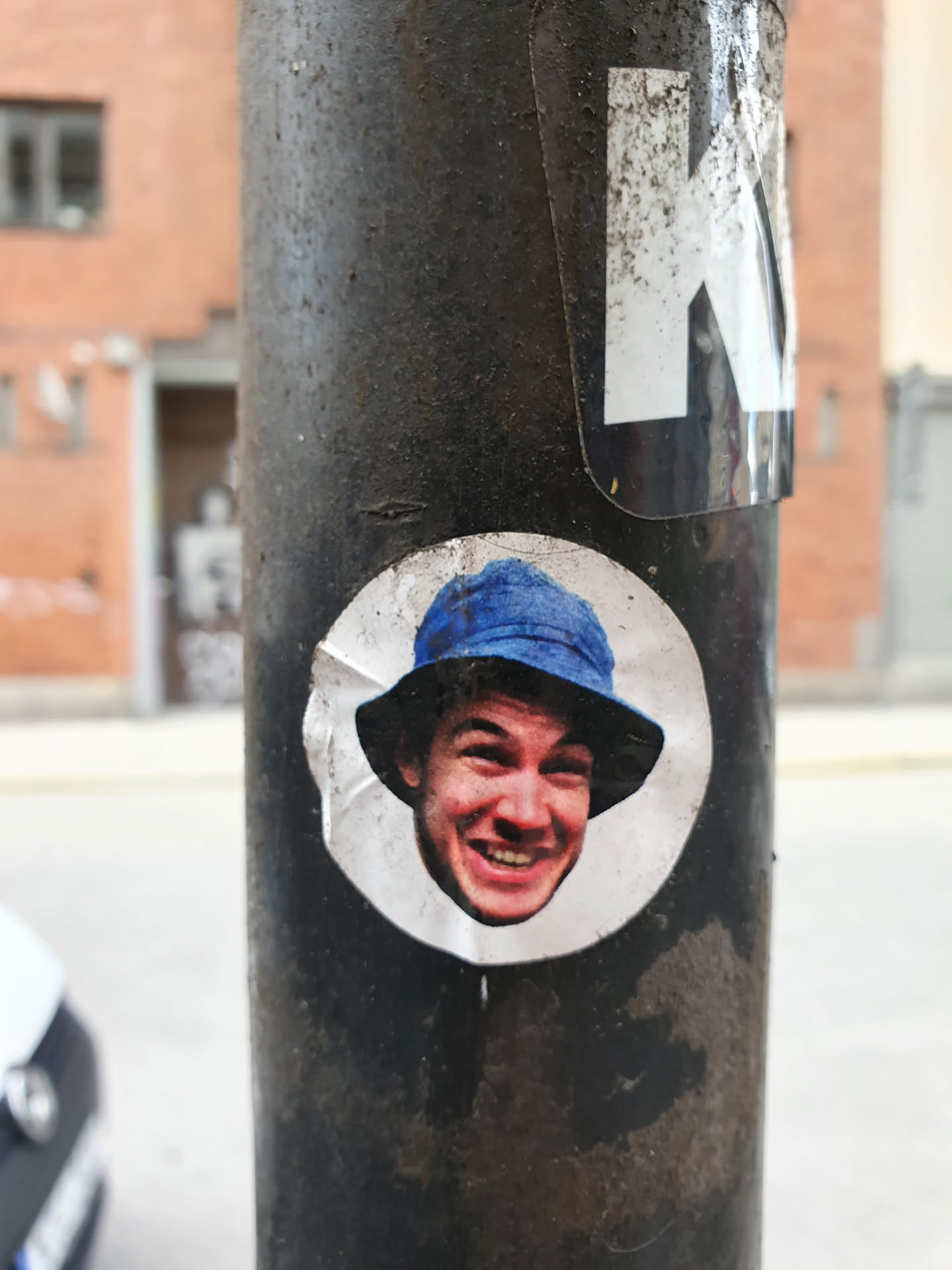 A round sticker featuring a close-up photo of a smiling man wearing a blue bucket hat. The sticker is affixed to a dark-colored pole outdoors.