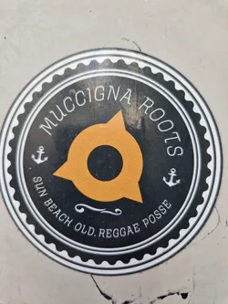 Round sticker with a black and white design. It features the text "MucciGNA Roots" on the top, and at the bottom it says "Sun Beach Old Reggae Posse". There is an orange compass rose-like figure in the center, and anchors on each side.