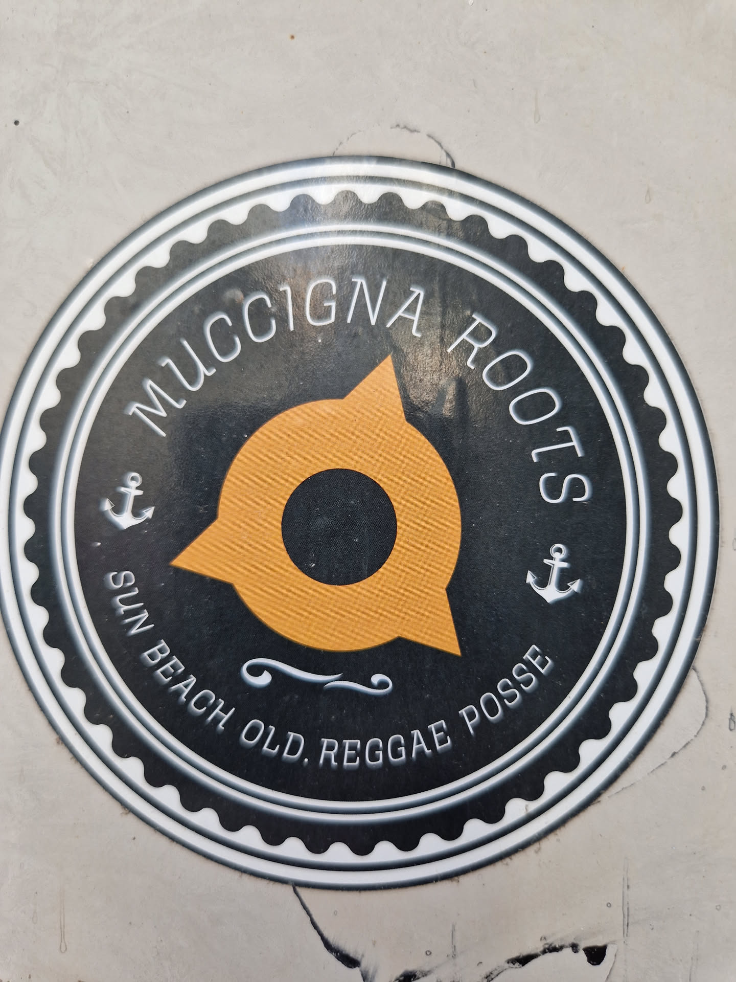 Round sticker with a black and white design. It features the text "MucciGNA Roots" on the top, and at the bottom it says "Sun Beach Old Reggae Posse". There is an orange compass rose-like figure in the center, and anchors on each side.