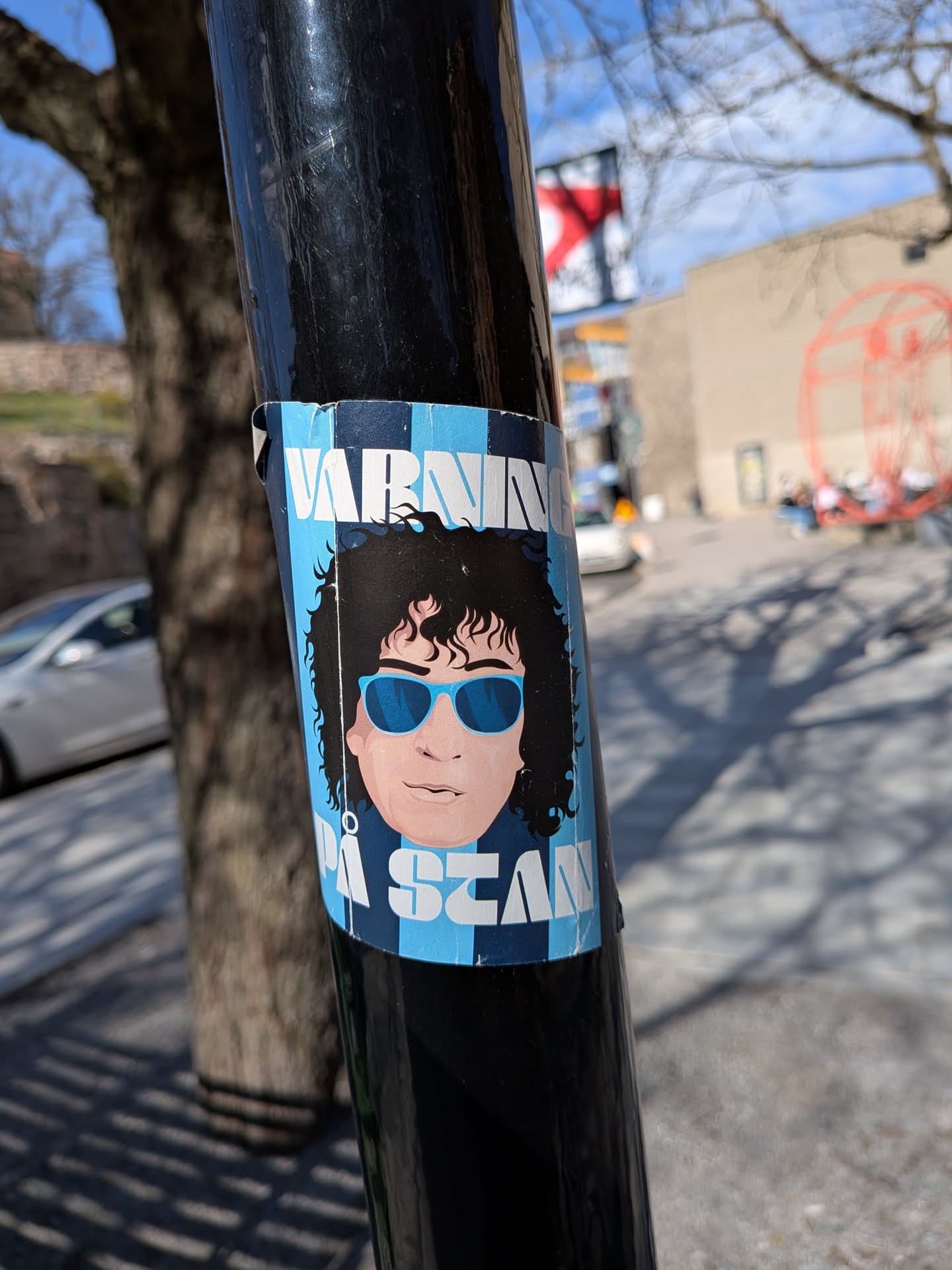 A rectangular sticker adhered to a black pole, featuring a stylized cartoon-like depiction of a person from the shoulders up. The character has dark, voluminous curly hair and is wearing bright blue sunglasses. Their skin tone is a light peach. The background of the sticker is composed of alternating light blue and dark blue vertical stripes, with a checkerboard pattern visible at the very top. White block letters are overlaid on this striped background. The sticker shows signs of wear, with slight peeling and creasing, particularly at the top and bottom edges, indicating it's a physical sticker exposed to the elements.