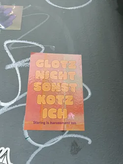 A sticker with German text that translates to "Don't stare or I'll vomit." It also includes the message "Staring is harassment too." The design features bubble-like letters in a peach color gradient on a peach background, along with a small pink illustration of what looks like a llama or alpaca.