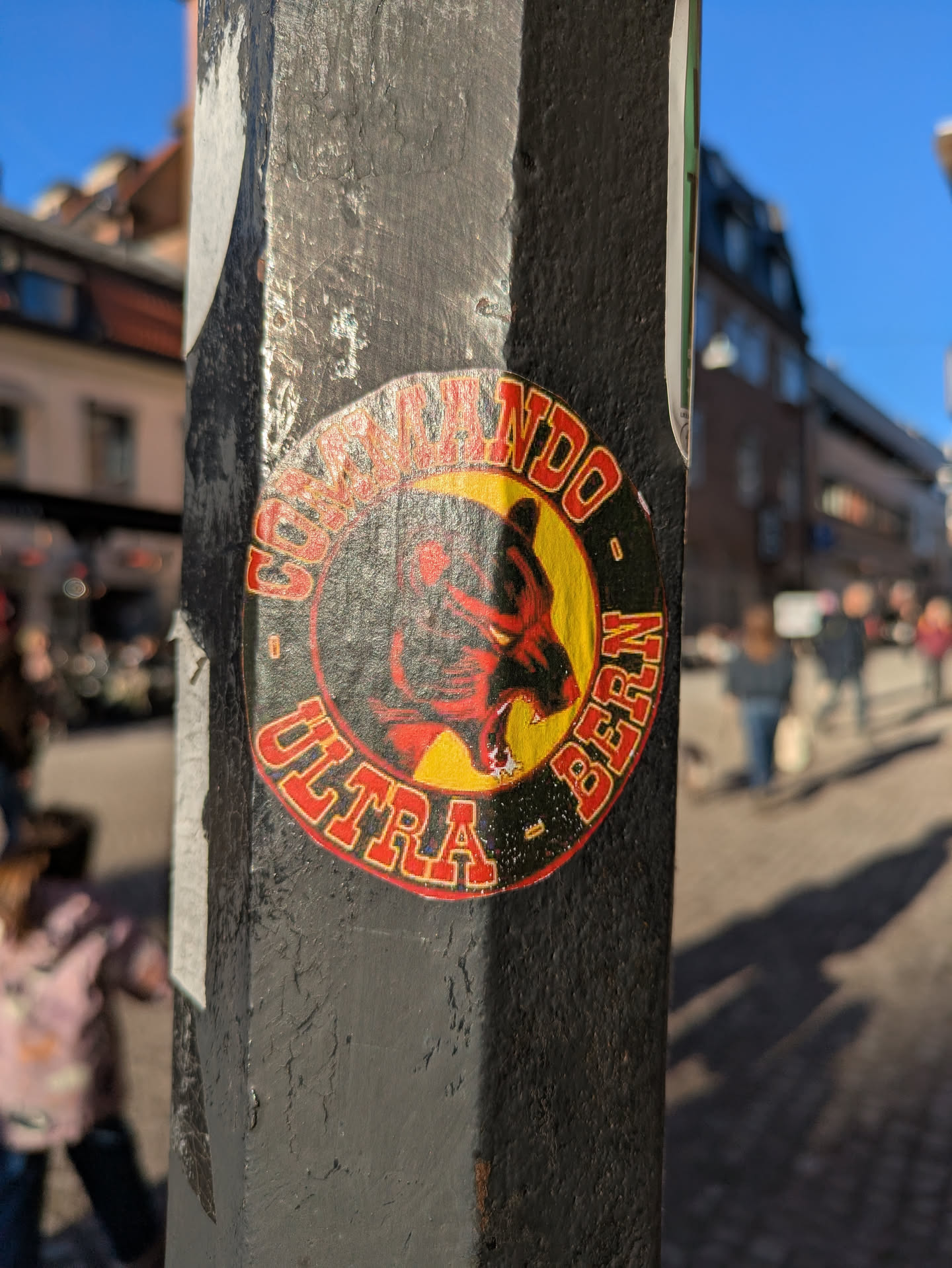 This sticker is on a pole. It features a bear logo with a crescent moon behind it, with the words "Commando Ultra Bern" encircling the image. The color scheme is primarily red, yellow, and black.