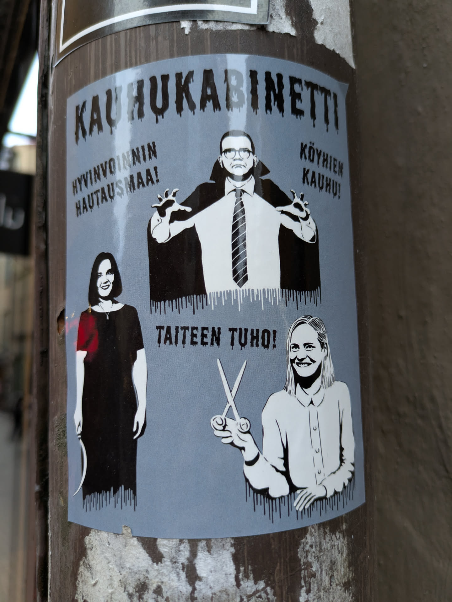 A sticker featuring a stylized illustration of three individuals.  A man in a Dracula-like cape is centrally positioned, flanked by a woman on the left and another woman on the right.  Text in Finnish surrounds the figures, suggesting a theme of horror or satire related to wellbeing, poverty, and art.