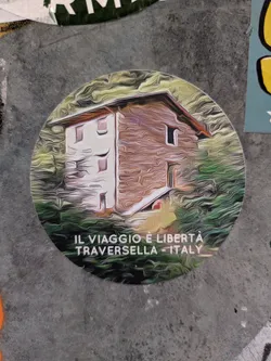 Round sticker featuring a stylized painting of a house in Traversella, Italy. The text at the bottom reads: "IL VIAGGIO È LIBERTÀ TRAVERSELLA - ITALY"