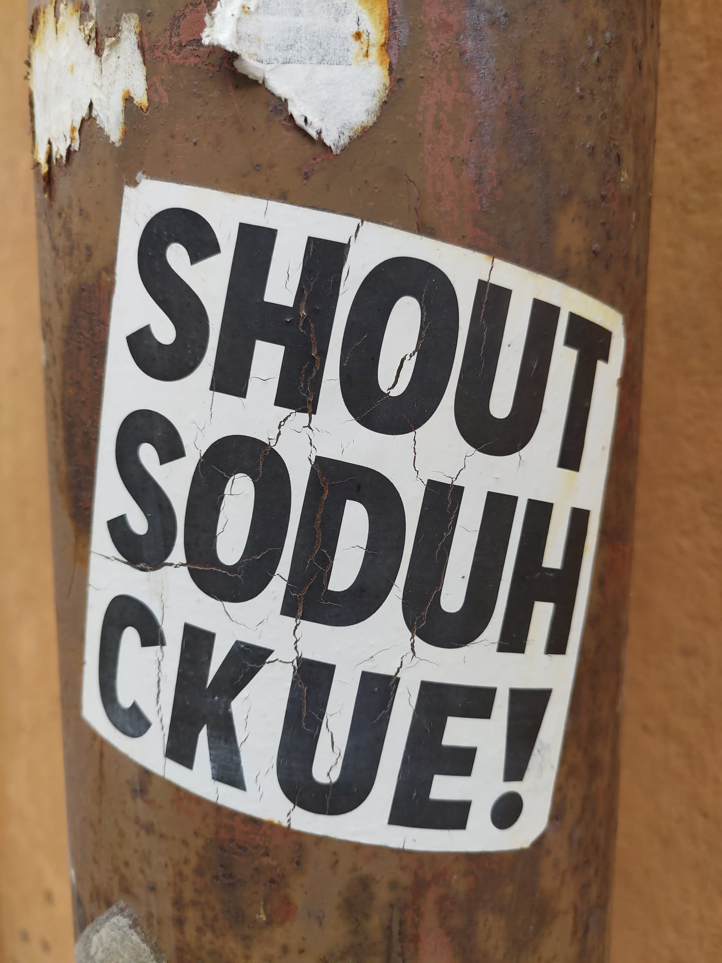 A worn sticker with the text "Shout Soduh CKUE!" on a rusty surface. The sticker shows signs of age and weathering, adding to its character. The bold, uppercase lettering is simple yet effective, conveying a clear message.