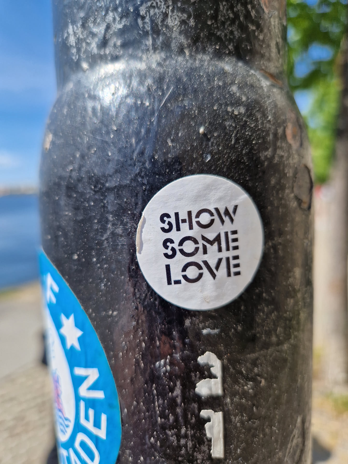 A round sticker on a dark pole. The sticker is white with black text that says SHOW SOME LOVE in a stylized font. 