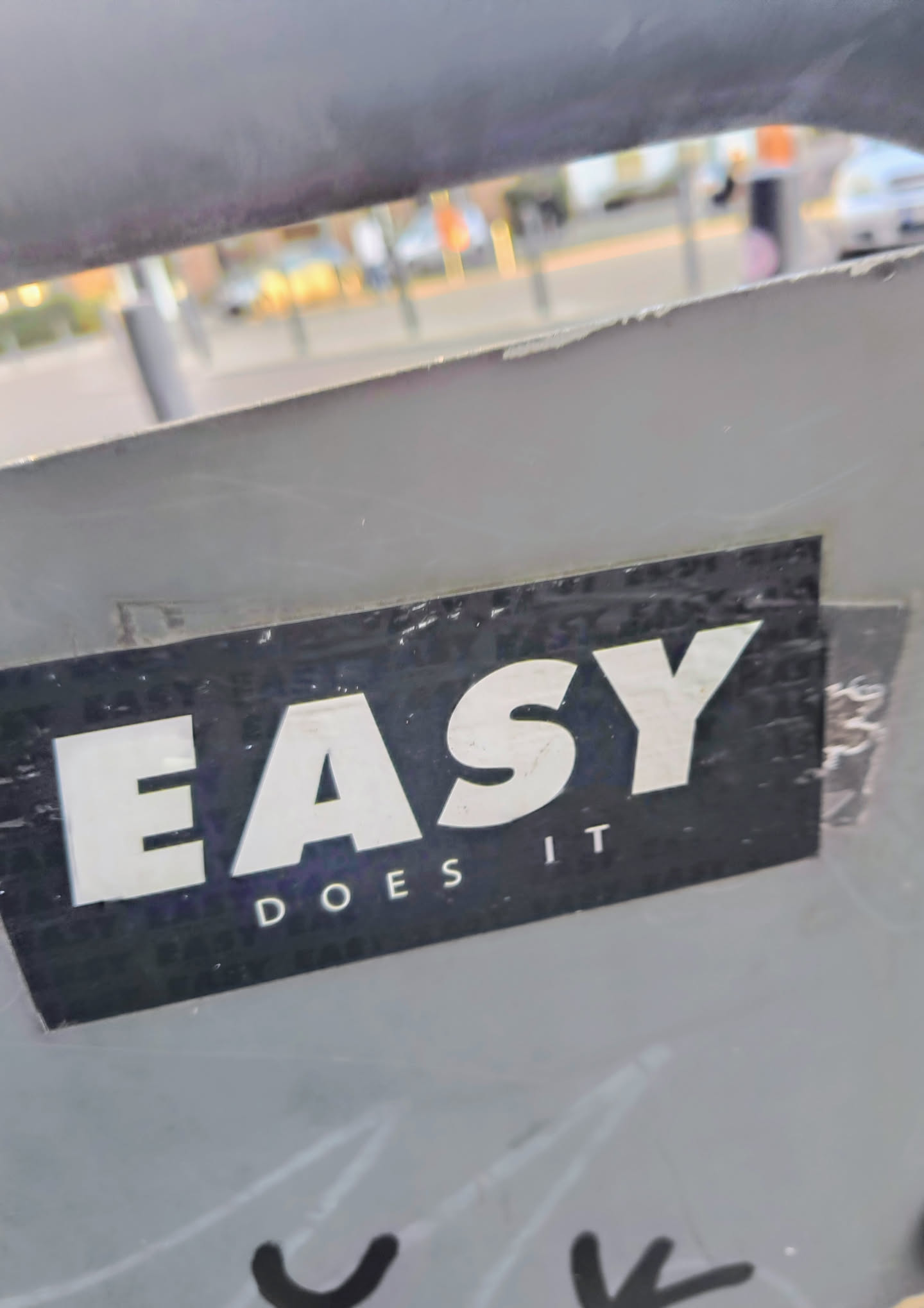 A black sticker with white text "EASY DOES IT" is adhered to a light gray, possibly metallic, surface. The sticker is rectangular, with the word "EASY" in large letters at the top and "DOES IT" in smaller letters beneath it. The sticker's corners are slightly worn, and it appears to be held in place with tape in some areas. The background is blurry, suggesting an urban outdoor setting.