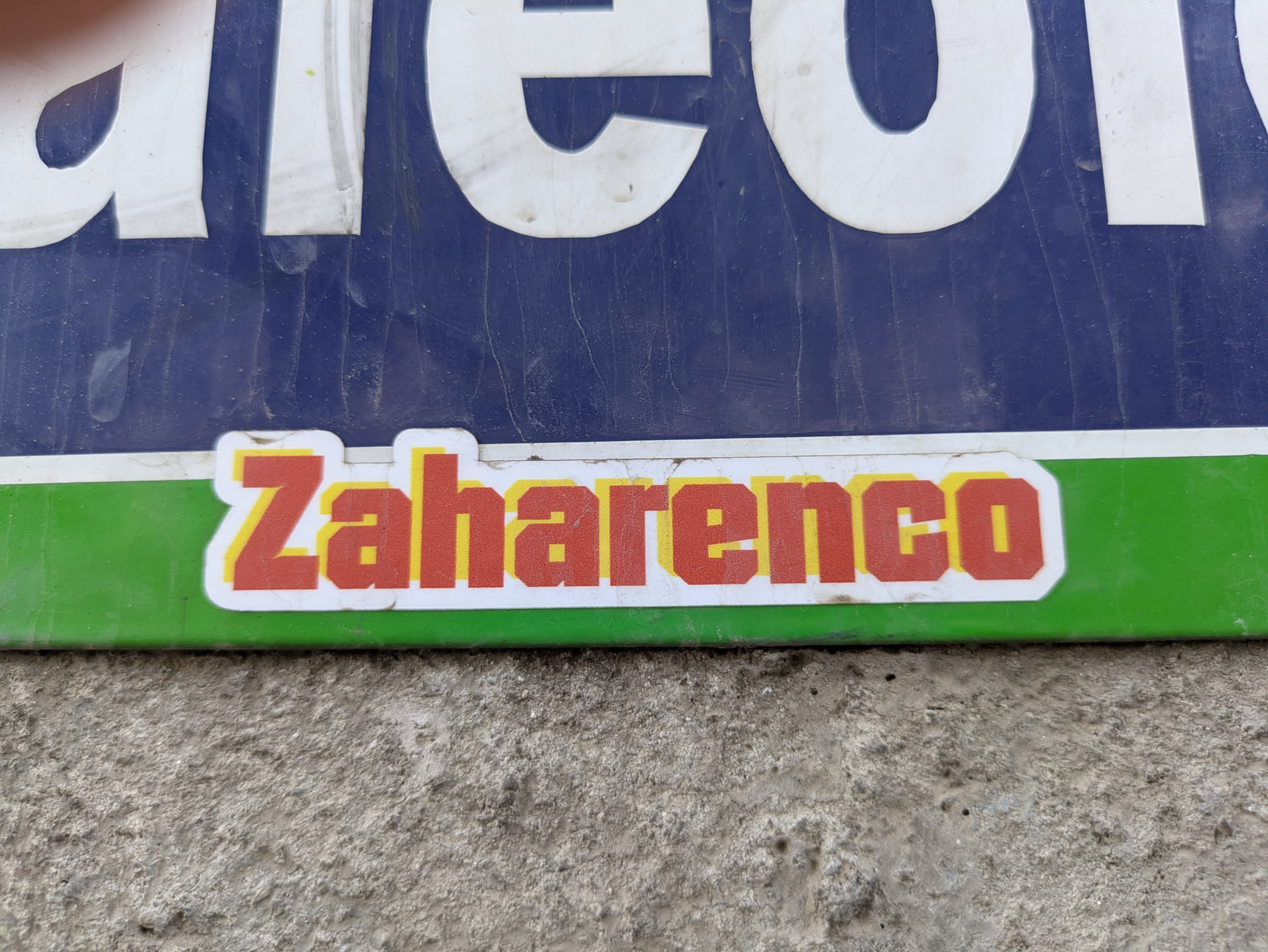 A sticker that says 'Zaharenco' in red with a yellow border on a white background. The sticker is placed on a green and blue sign.