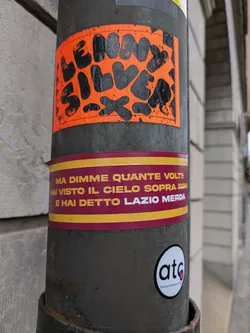 An orange sticker with the name Lenny Silvera written in a stylized font. The sticker has a distressed look, giving it a vintage feel.  Below it is another sticker with Italian text that roughly translates to: "But tell me how many times you've seen the sky above Rome and said Lazio Merda."  There is a third sticker that says 'atg'