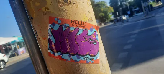 A rectangular sticker with rounded corners, affixed to a weathered, peeling yellow pole. The top portion features a solid orange background with black text reading "HELLO MY CREW IS". The main design consists of large, stylized, bubbly purple letters, possibly spelling "YUGSS", outlined in black with subtle orange highlights. These letters are set against a background of various blue and light blue bubble or cloud-like shapes, some accented with darker blue outlines and small orange dots. A small, illegible black signature or tag is visible in the bottom right corner of the main design. The overall aesthetic is reminiscent of graffiti and street art.