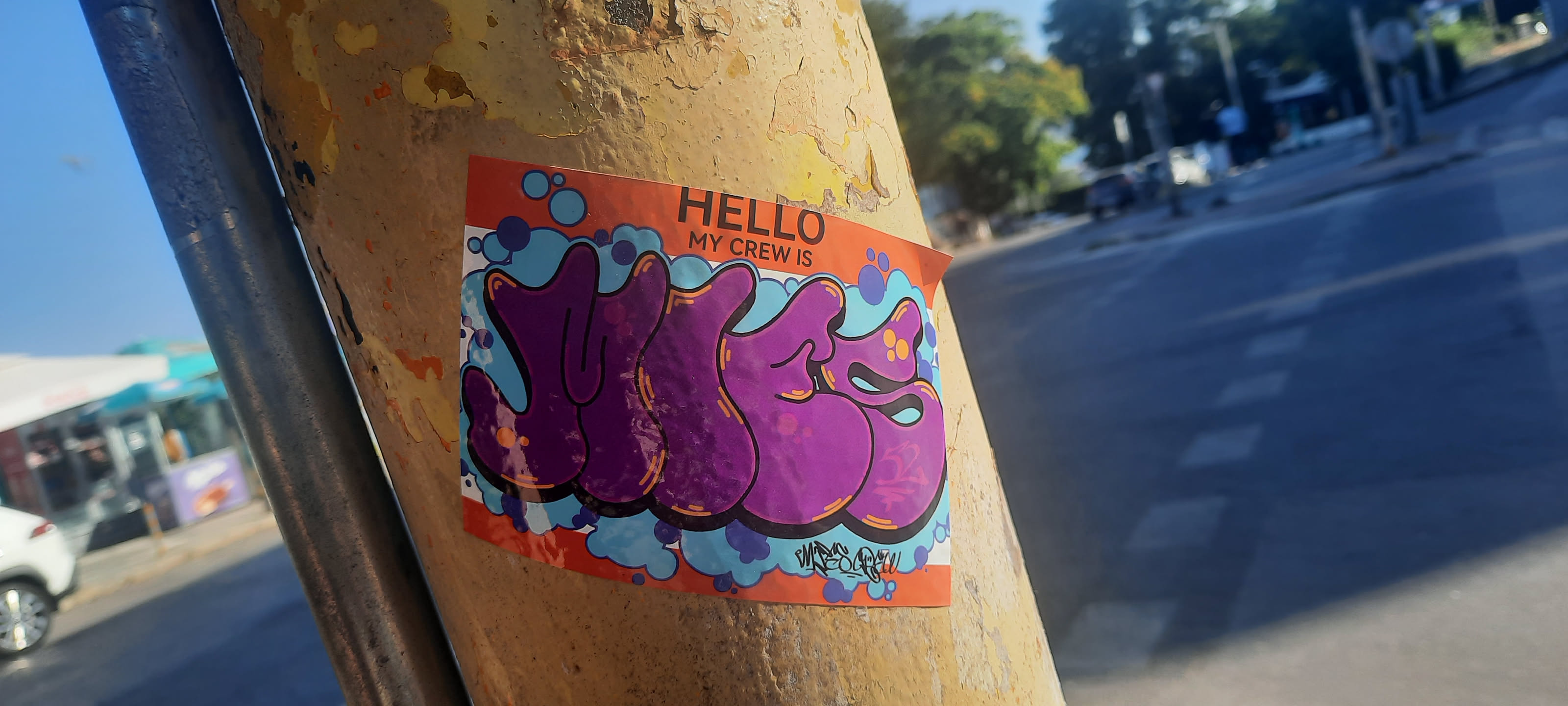 A rectangular sticker with rounded corners, affixed to a weathered, peeling yellow pole. The top portion features a solid orange background with black text reading "HELLO MY CREW IS". The main design consists of large, stylized, bubbly purple letters, possibly spelling "YUGSS", outlined in black with subtle orange highlights. These letters are set against a background of various blue and light blue bubble or cloud-like shapes, some accented with darker blue outlines and small orange dots. A small, illegible black signature or tag is visible in the bottom right corner of the main design. The overall aesthetic is reminiscent of graffiti and street art.