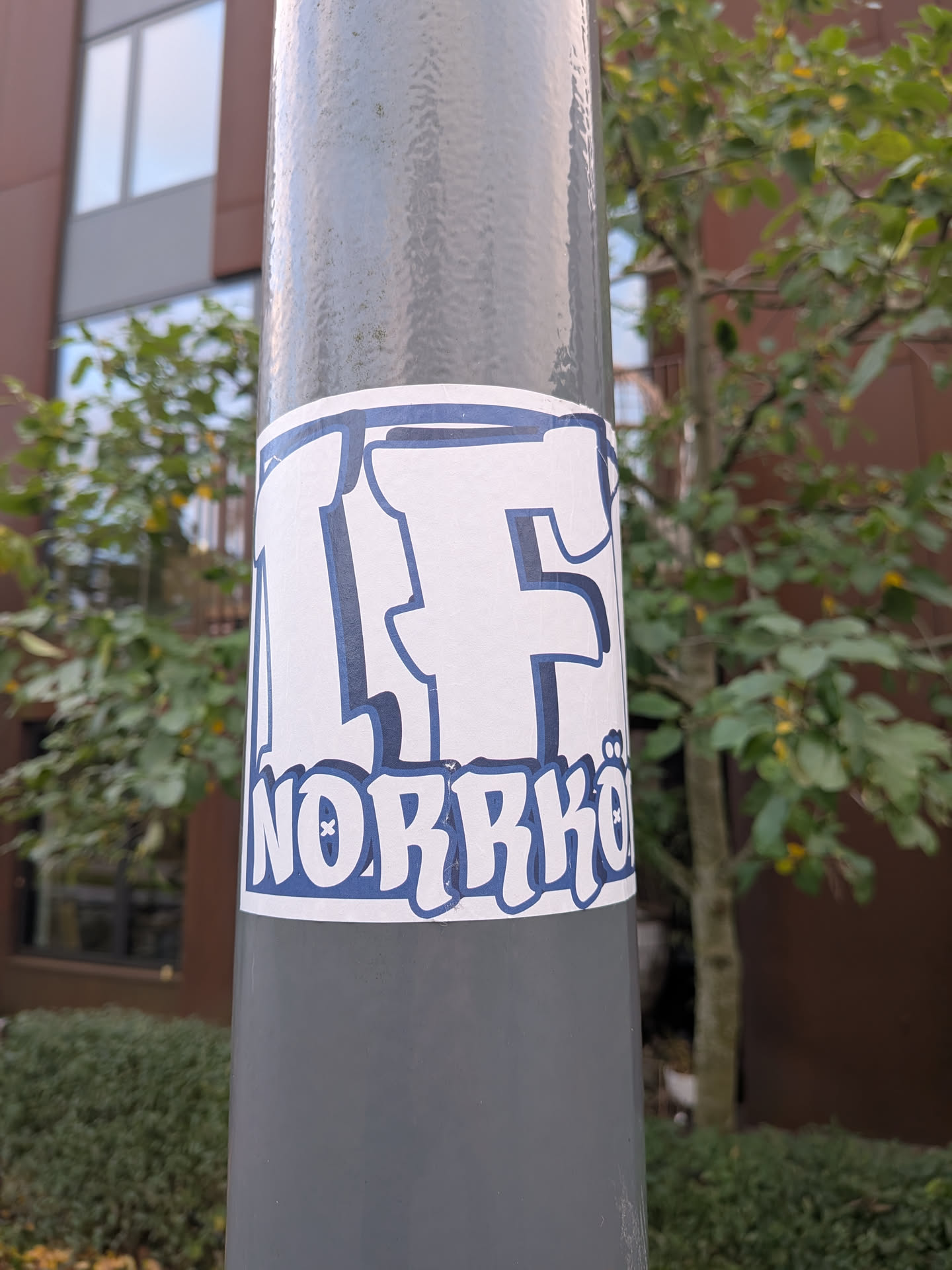 A rectangular adhesive sticker wrapped around a cylindrical grey metal post. It features bold, graffiti-inspired lettering in white with a dark blue outline and shadow. The top half shows large stylized letters reading 'IF', while the bottom line displays 'NORRKÖ' with small decorative dots forming an umlaut above the 'O', representing the Swedish football club IFK Norrköping.