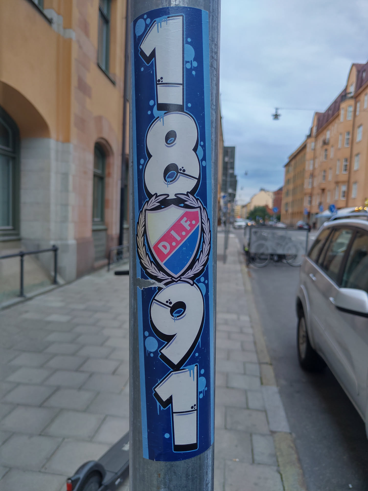 A vertical sticker featuring the numbers 1891 in a stylized graffiti font, with a central emblem of D.I.F (presumably a football club). The sticker is applied to a metal pole on a city street.