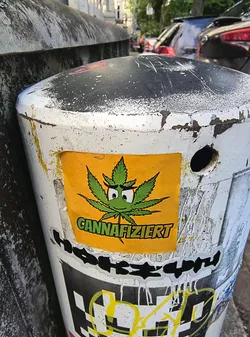 A rectangular yellow sticker featuring a cartoon cannabis leaf with a smiling face and the word 'CANNAFIZIERT' is attached to a white and black bollard on a city street. The sticker is colorful and stands out against the aged bollard.
