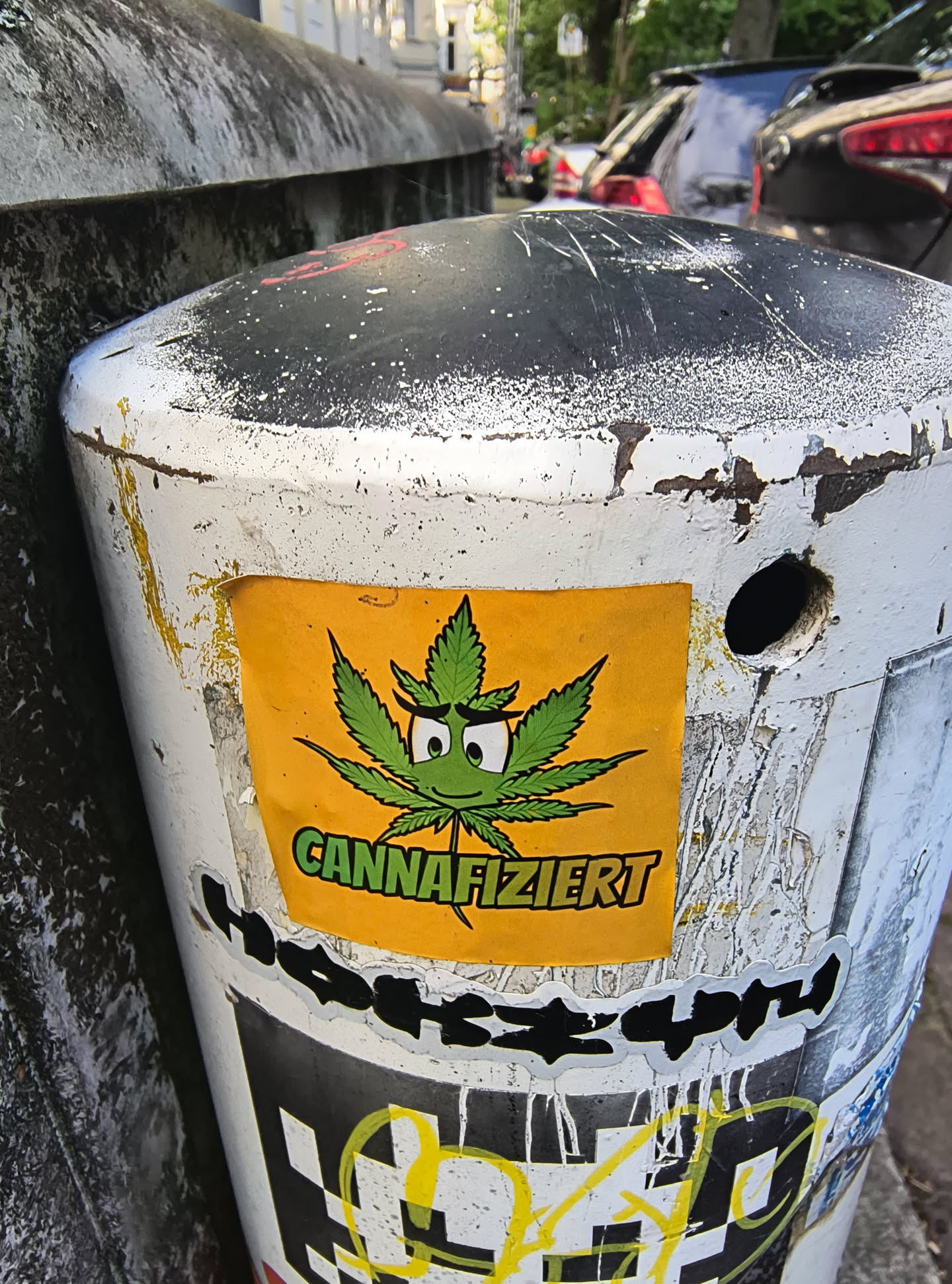 A rectangular yellow sticker featuring a cartoon cannabis leaf with a smiling face and the word 'CANNAFIZIERT' is attached to a white and black bollard on a city street. The sticker is colorful and stands out against the aged bollard.