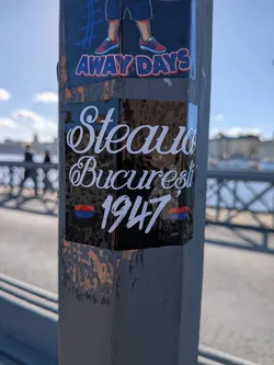 Street sticker A rectangular black glossy sticker adhered to a wooden post outdoors. The sticker features white cursive script typography spelling 'Steaua' on the first line and 'București' on the second line. Underneath, the founding year '1947' is written in a stylized white font, flanked on either side by small horizontal red and blue stripes representing the team's iconic colors.
