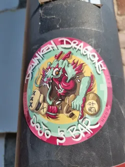 A round sticker featuring a cartoon illustration of a green dragon wearing boxing gloves and holding a sake bottle. The text on the sticker says "Drunken Dragon Dojo 5 Bar". The sticker has a pink and yellow color scheme.