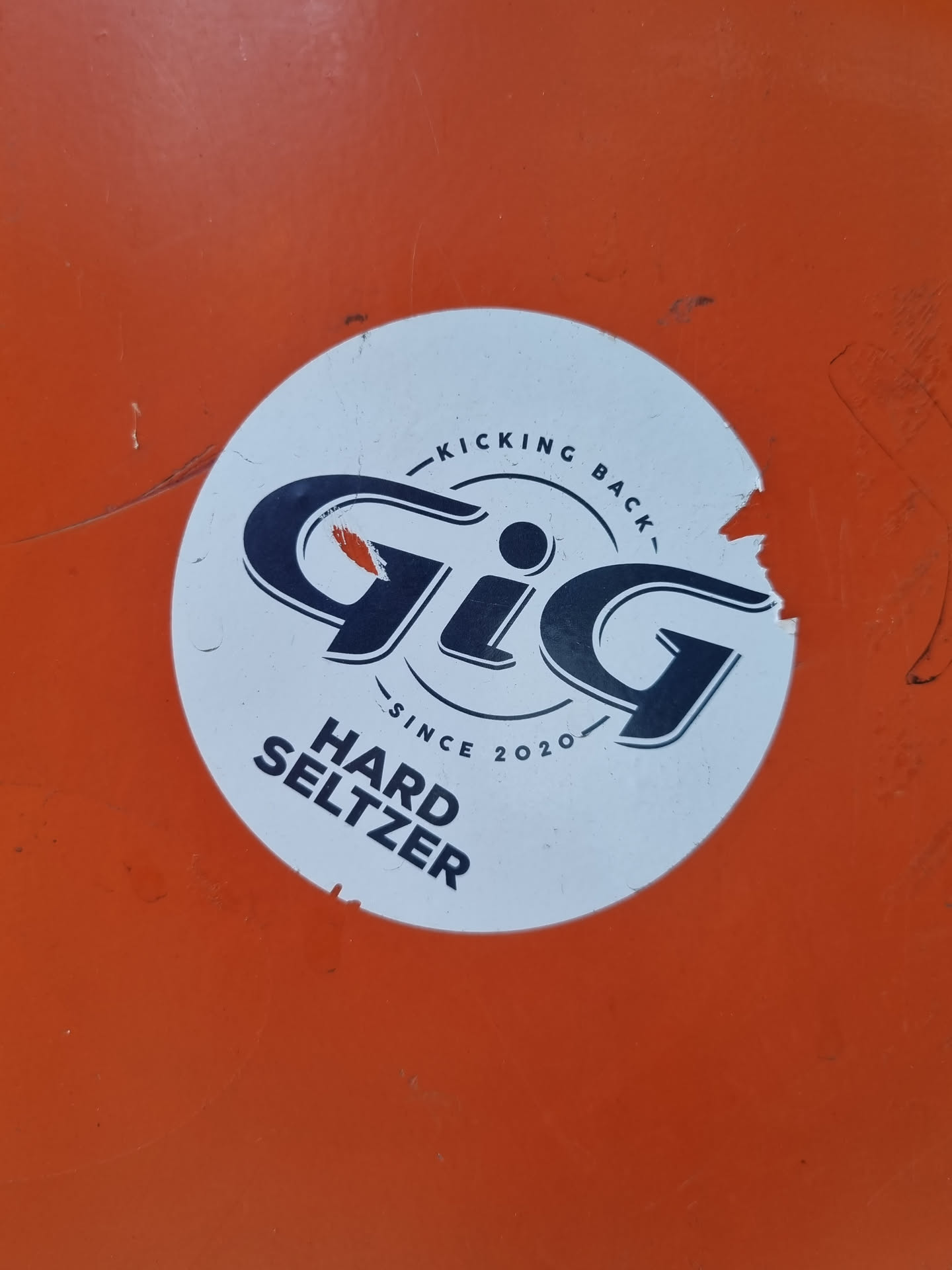A white circular sticker with the Gig Hard Seltzer logo. The logo features the word "Gig" in a stylized font, along with the text "Kicking Back" and "Since 2020". The sticker is slightly torn at the edges and is affixed to an orange surface.