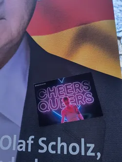 A sticker featuring the text 'Cheers Queers' in neon style with an image of a person. The sticker is placed on a dark surface with the text 'Olaf Scholz' visible in the background.