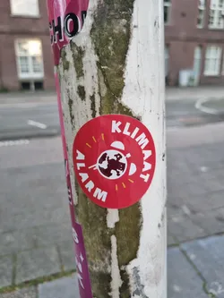 A red circular sticker with the text "KLIMAAT ALARM" (Climate Alarm in Dutch) and a graphic of the Earth.  The sticker is affixed to a weathered pole.