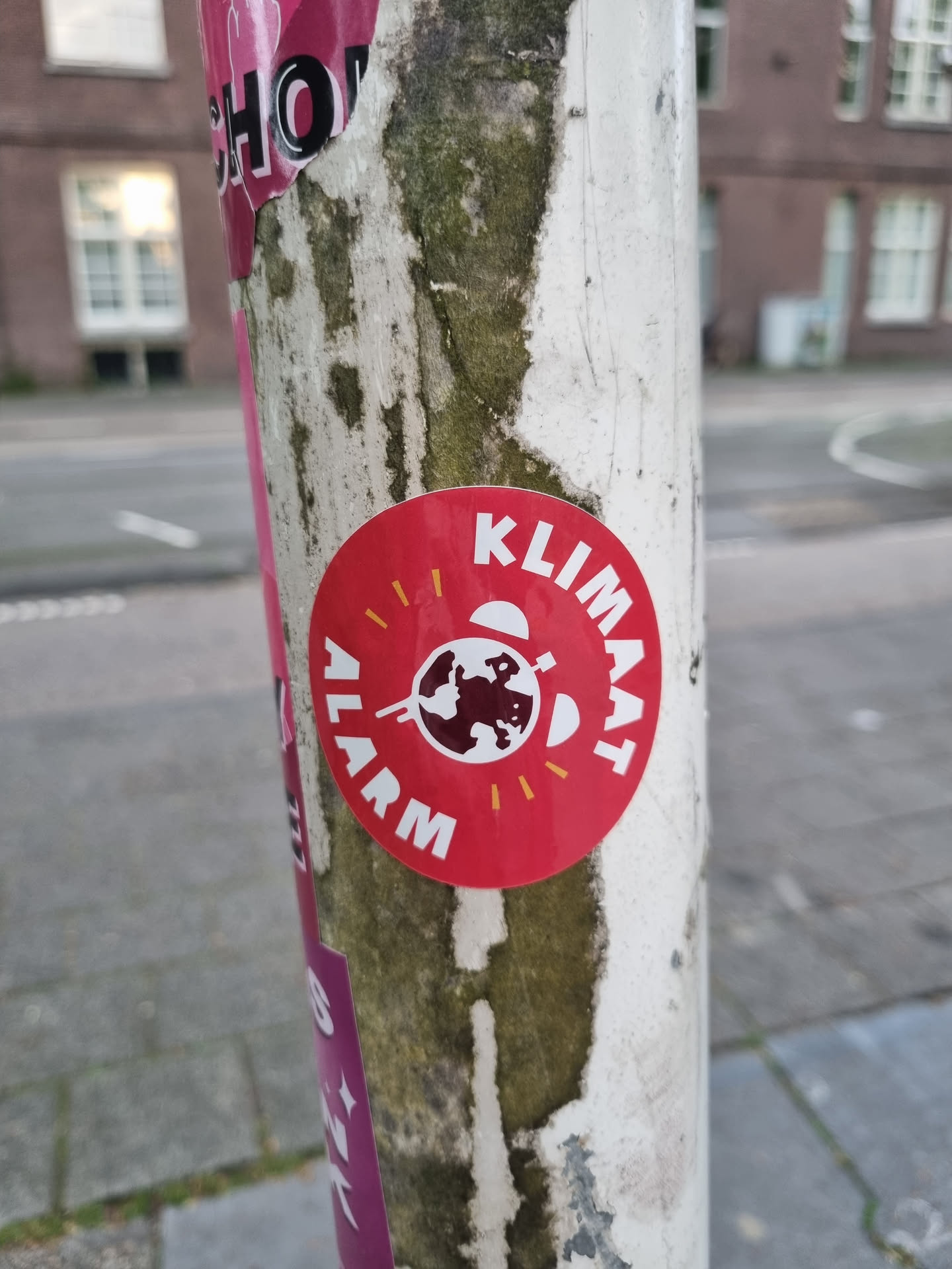 A red circular sticker with the text "KLIMAAT ALARM" (Climate Alarm in Dutch) and a graphic of the Earth.  The sticker is affixed to a weathered pole.