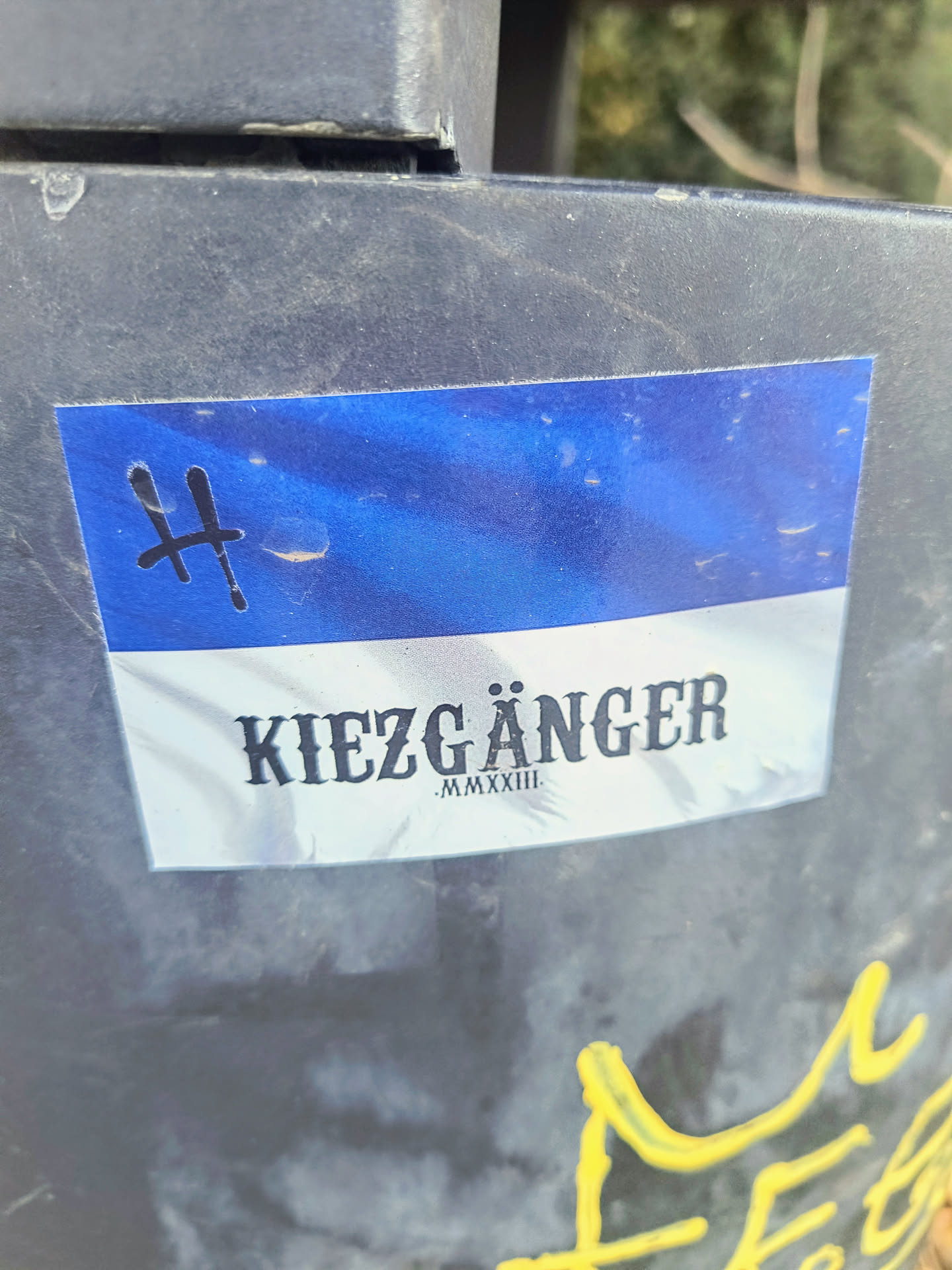 A rectangular sticker with a blue top and white bottom is affixed to a trash can. The top portion of the sticker is a mottled blue, while the bottom is white with the text "KIEZGÄNGER" and below it "MMXXIII". To the left, there's a symbol resembling an 'H' or a stylized cross.
