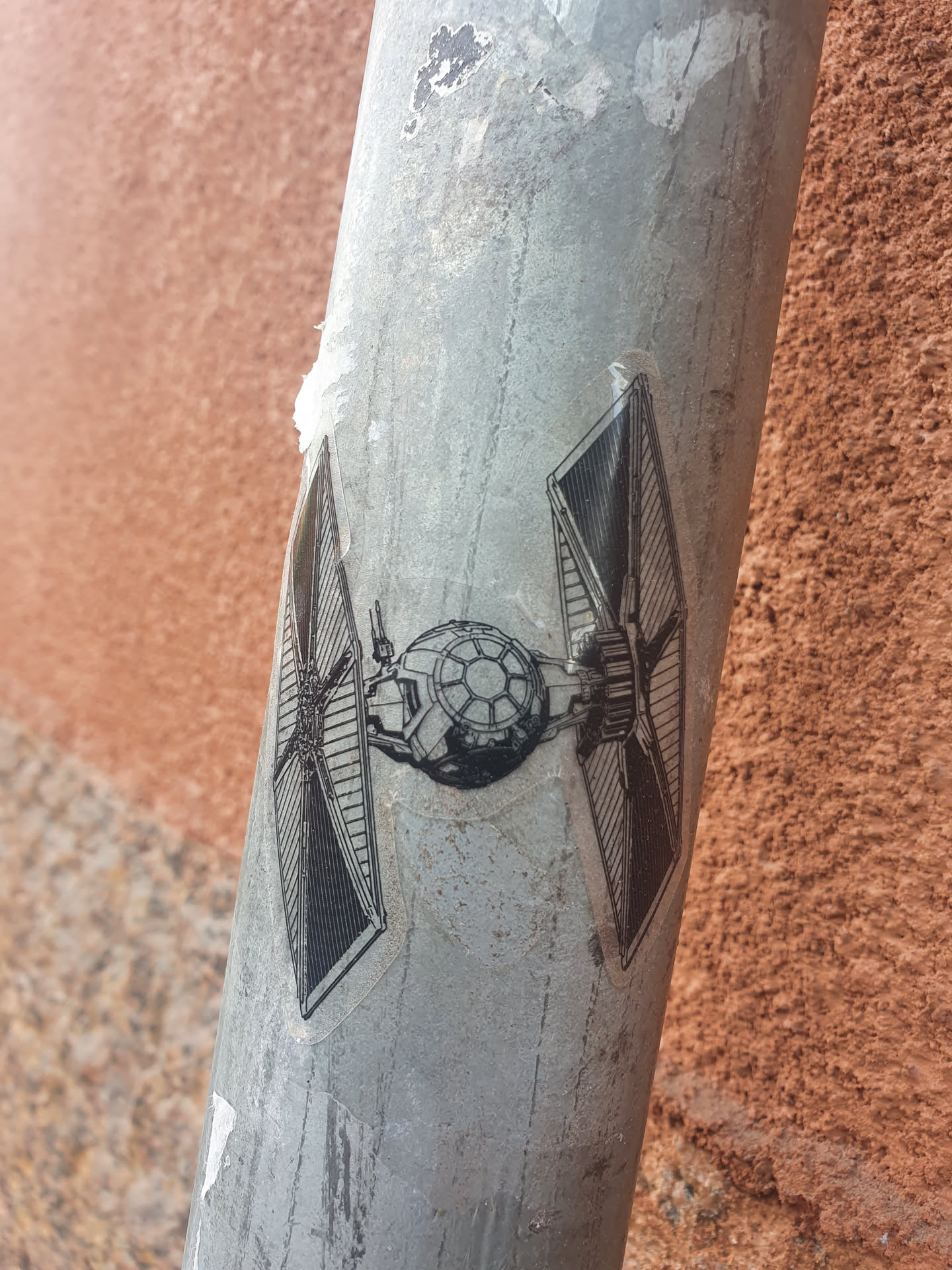 A grayscale sticker depicting a TIE Fighter from Star Wars is affixed to a weathered metal pole. The sticker shows some wear, suggesting it has been exposed to the elements for some time. The background is a textured, reddish-brown wall.