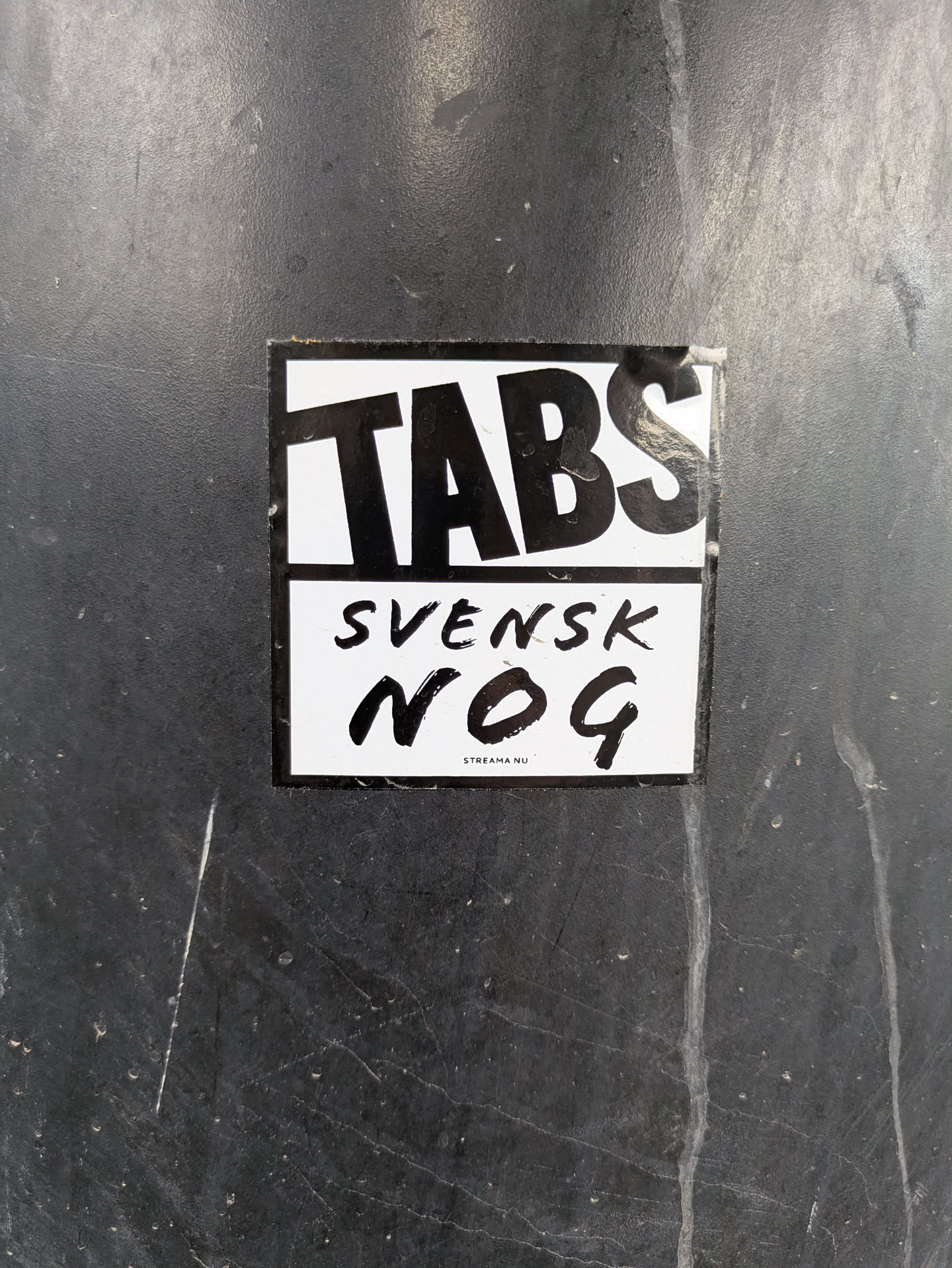 The sticker is rectangular with a white background and black text. The top section prominently features the word 'TABS' in large, bold, sans-serif letters. Below 'TABS', in a slightly smaller font, is the word 'SVENSK', followed by 'NOG' in a large, distinct, handwritten or brush stroke style font. At the very bottom, in small, uppercase sans-serif text, it reads 'STREAMA NU'. The sticker itself appears slightly worn around the edges and is affixed to a dark, possibly metallic or plastic, textured surface with some white scuff marks.