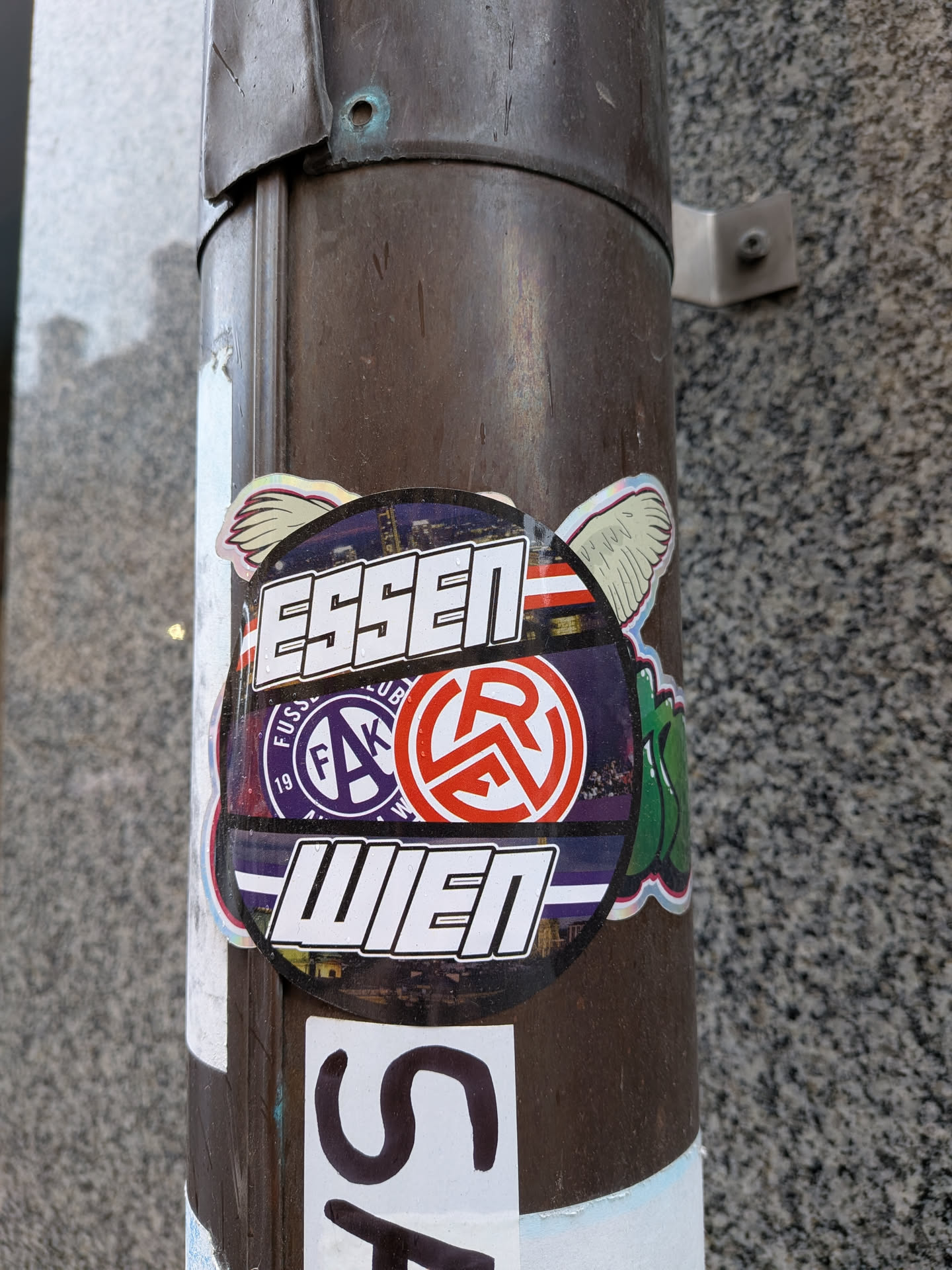 The sticker is predominantly circular with white, feathered wings extending from its upper sides, giving it a dynamic appearance. The main body features a dark, textured background, possibly depicting a cityscape or stadium lights, overlaid with subtle horizontal red and blue stripes. Bold white block letters with a black outline spell 'ESSEN' across the top and 'WIEN' across the bottom. In the center, there are two circular football club logos: on the left, a purple logo with 'FUSSBALL CLUB', 'FAK', and '1911' in white text; on the right, a red logo with a stylized white 'R' or 'RA' symbol. The sticker has a glossy finish and shows minor wear, indicating it's a physical sticker adhered to a metal pole.